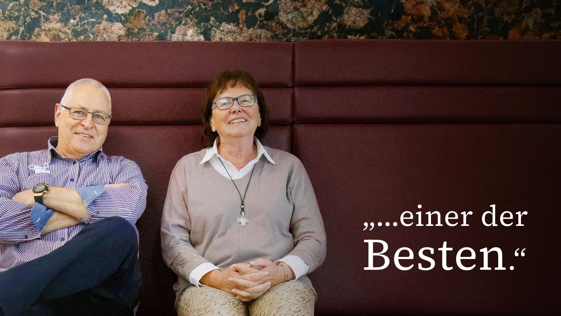 Zwei Personen sitzen auf roter Bank mit Schriftzug „… einer der Besten.“